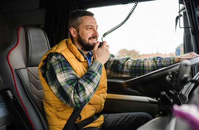 A driver of a fleet is seen in his truck, smiling and talking into a CB radio while driving.